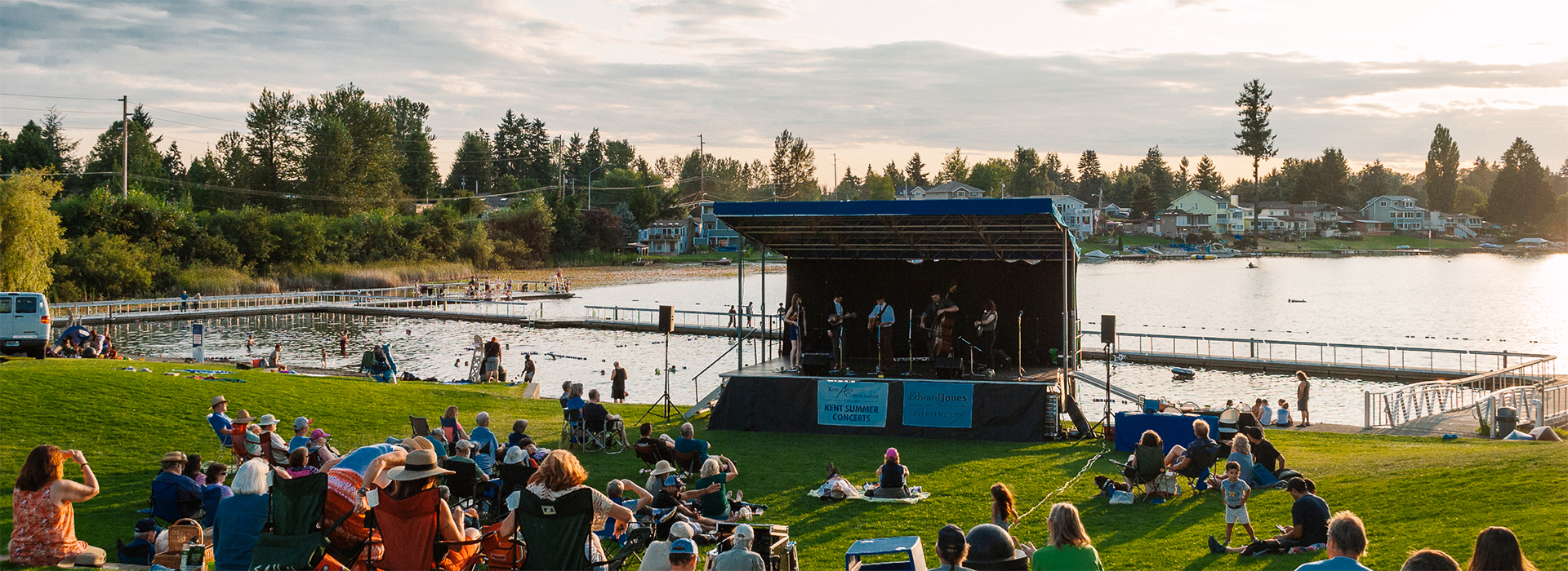 A crowd is gathered on the lawn facing the summer concert stage by the lake shore.