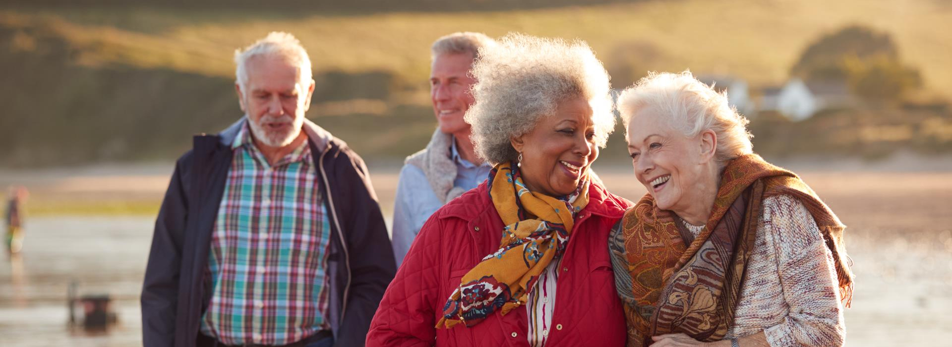 A group of four senior friends laugh as they walk along a beach.