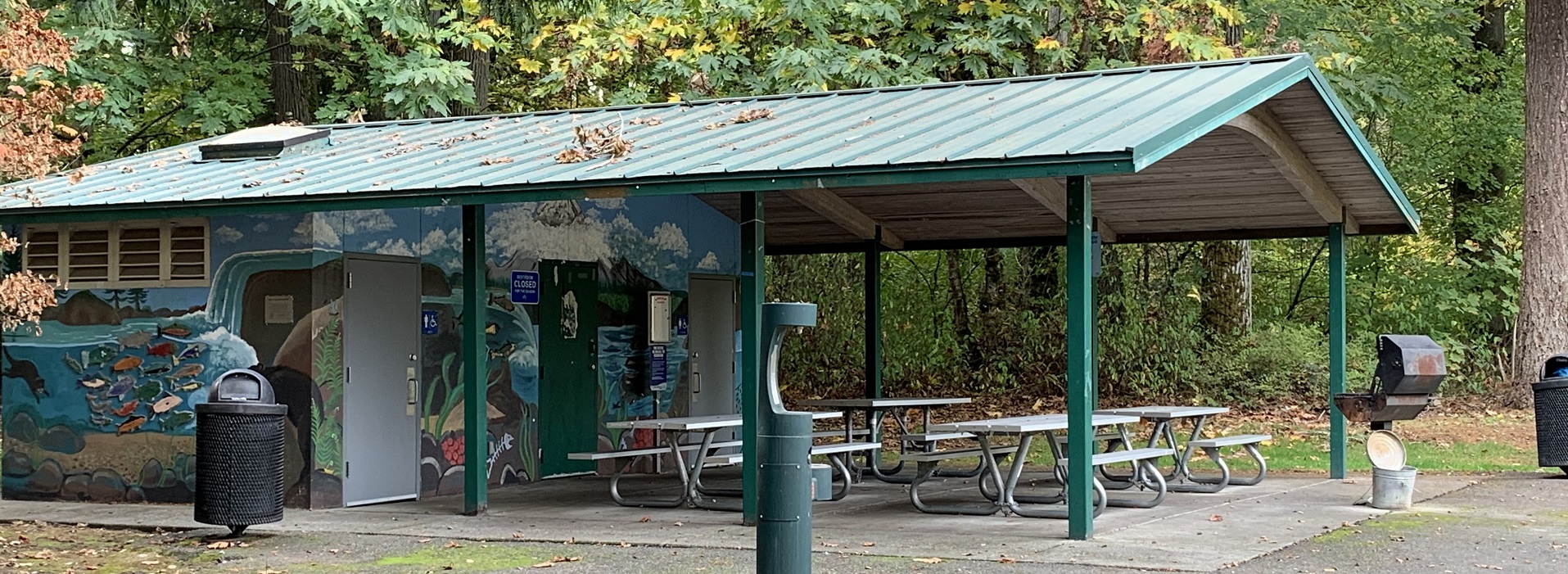 Covered picnic shelter with metal tables, restrooms and a wall mural in front of leafy trees