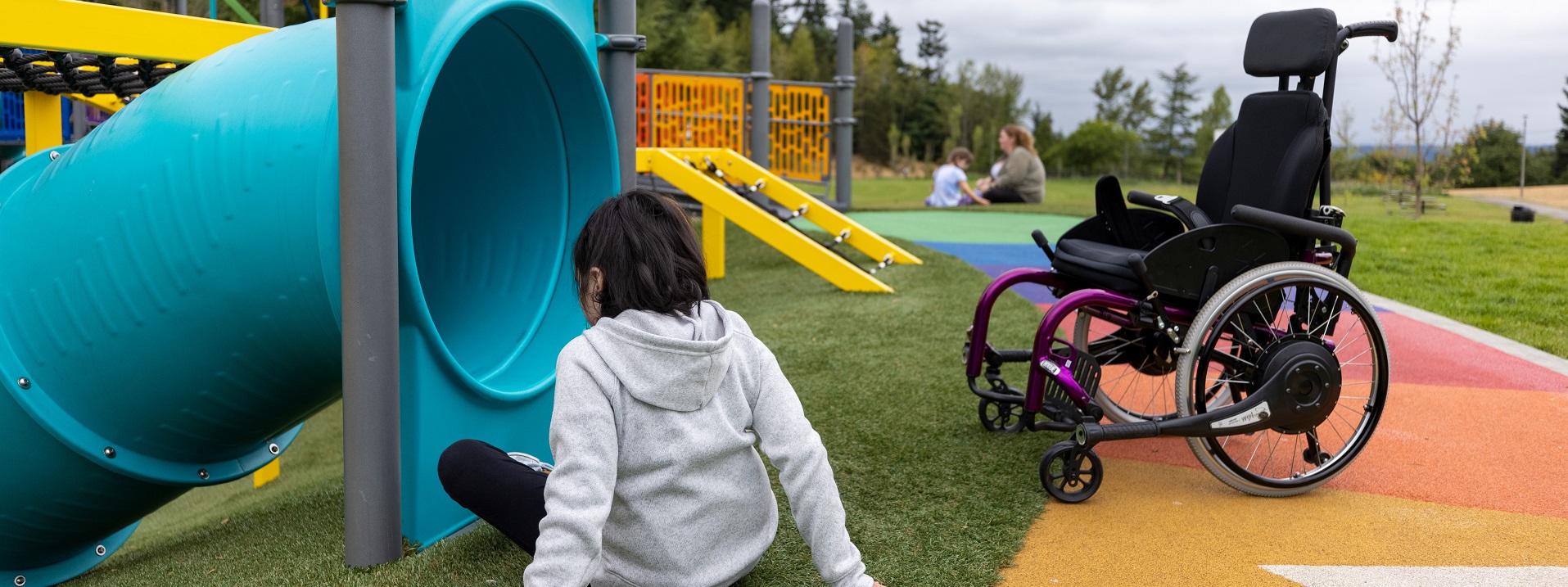 Young person wearing a grey hoodie sits on the grass by a blue slide tube and an empty wheelchair on the nearby colorful path