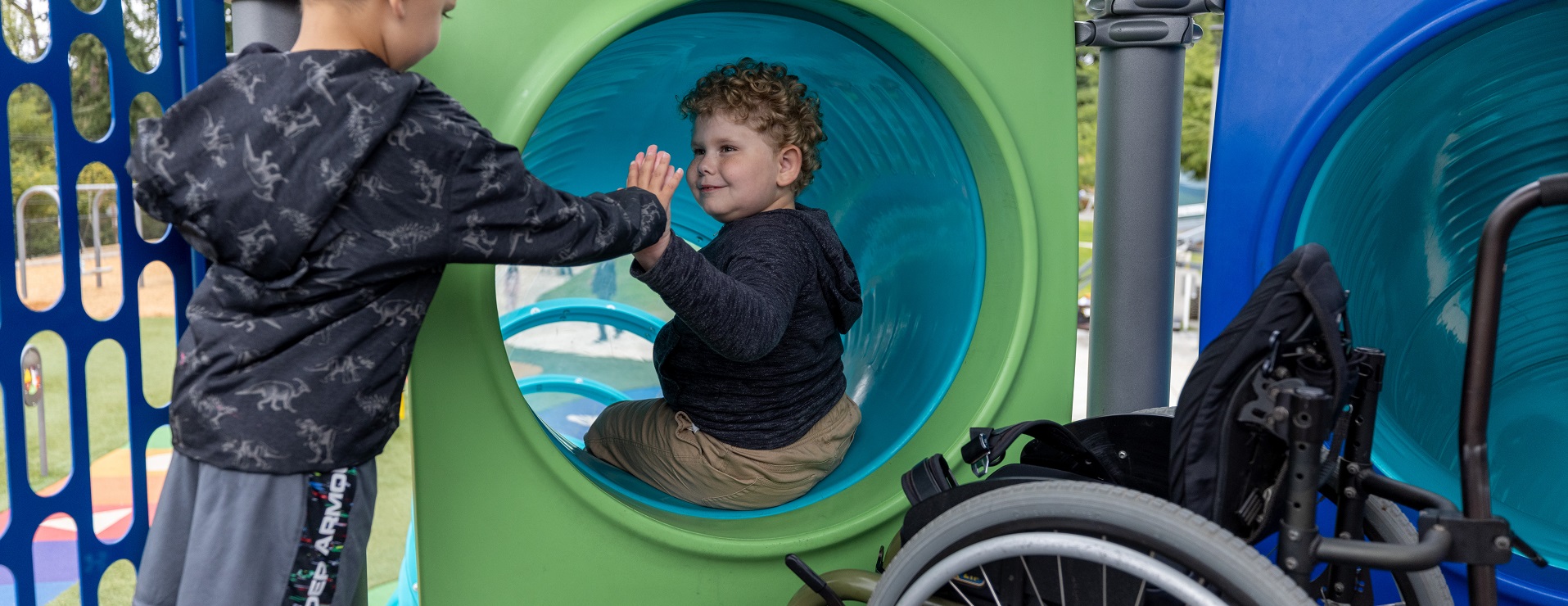 A child with curly hair sitting at the top of a slide gives a high-five to a child standing nearby while an unoccupied wheelchair sits by them